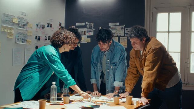 Creative crew of four young filmmakers standing around table, reviewing and discussing scene layouts and location photos during meeting in studio