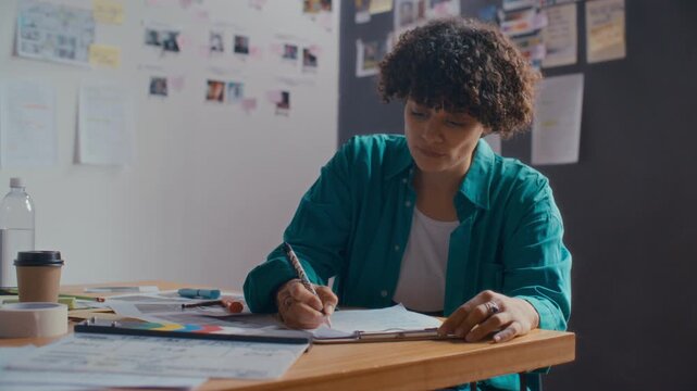Concentrated female screenwriter with curly hair sitting at desk in creative studio, writing script for film production and analyzing her notes
