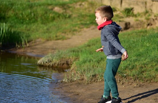 Young boy standing on a sandy lakeshore holding a stone and preparing to throw it into the water. Candid outdoor childhood moment of play, exploration and fun in nature on a sunny day.
