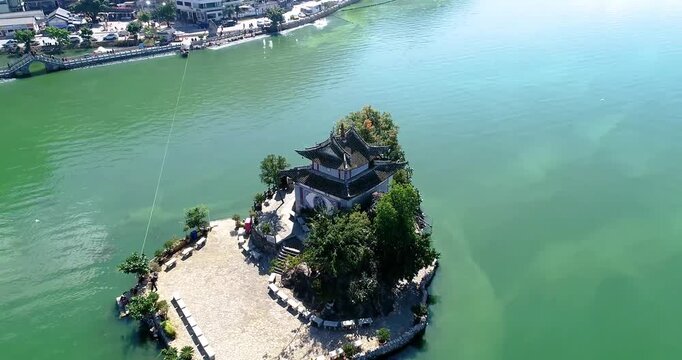 Aerial drone view of a traditional Chinese pavilion on a small island in Erhai Lake, Dali, with turquoise water, stone walkways, and surrounding shoreline village