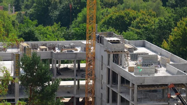 Construction of a modern concrete building involves heavy machinery. A tall yellow tower crane stands next to the monolithic framework structure. Workers install shuttering panels on the upper floor.