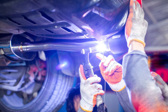 Selective focus to hands of auto mechanic Argon TIG wearing protective gloves performs precision welding on a car exhaust muffler pipe underneath the vehicle chassis in a garage repair workshop.