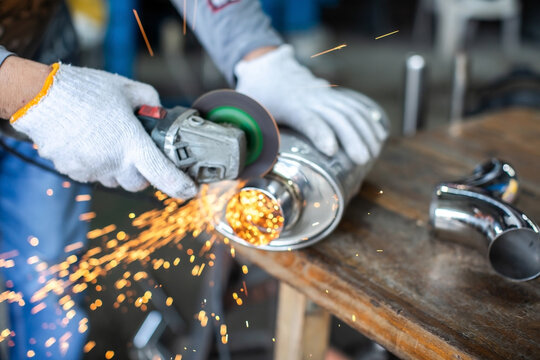 Blurry picture and bokeh of skilled worker wearing safety gloves uses an electric angle grinder to cut and smooth a metallic motorcycle exhaust component on a wooden workbench, creating bright sparks.