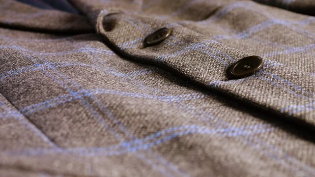 A sharp close-up of a brown wool tweed blazer with a blue plaid pattern and dark buttons, highlighting the high-quality fabric texture and classic masculine tailoring.