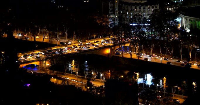 Cars crowd an illuminated bridge over the dark Mtkvari River in Tbilisi at night while streetlights and vehicle headlights reflect on the water beside a riverside road