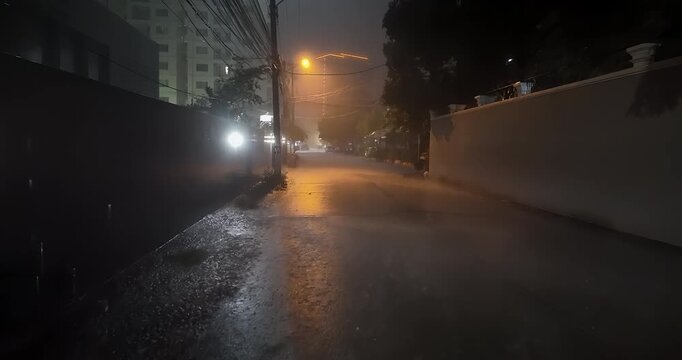 Walking on a small street while a motorcyclist rides through a rain drenched storm at night, followed by a bicycle traveling under the glowing haze of streetlights.