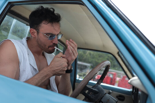 Cinematic horizontal shot of a young Caucasian man lighting a cigarette in a vintage automobile.