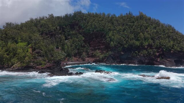 Aerial view of Kaihalulu Beach on Maui in the Hawaii island chain where turquoise ocean waves crash over black lava rocks below red volcanic cliffs covered with dense green tropical forest.