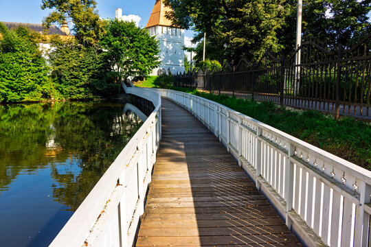A scenic wooden pedestrian bridge leading towards the ancient white walls and towers of St. Pafnutius Borovsky Monastery, surrounded by lush green trees and a calm water pond in Borovsk, Russia