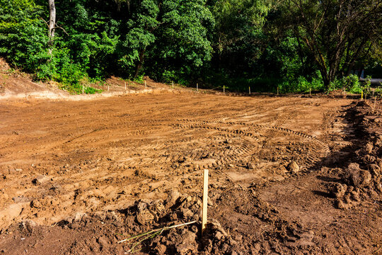 Freshly cleared construction site in a park featuring raw soil, deep tire tracks from heavy machinery, and survey stakes marking the perimeter for landscaping development