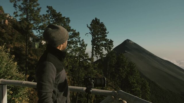 A photographer in warm clothing waits with a camera on a scenic viewpoint overlooking Fuego volcano, dawn sunlight casting long shadows across lush forested mountain slopes in Guatemala.