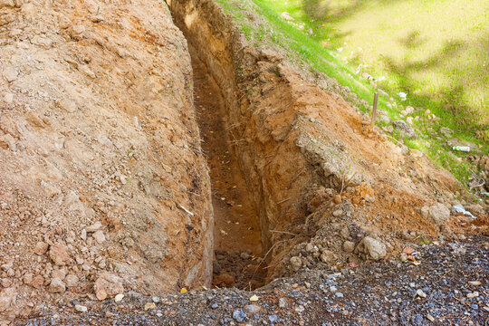 Deep excavated trench in clay soil for utility pipe installation showing raw ground layers at construction site on a sunny day with green grass along the edges