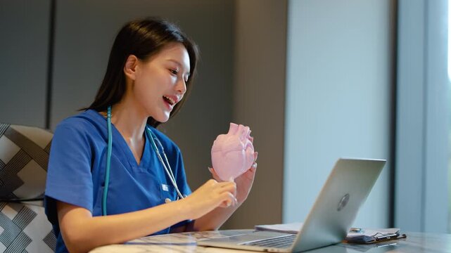 Smiling Asian female doctor in blue scrubs using a laptop for a telehealth video call, explaining cardiology concepts with a pink anatomical heart model to an online patient in a modern clinic.