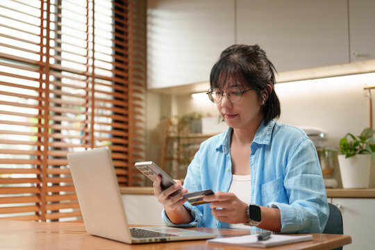 Asian Woman uses a smartphone and credit card with a laptop for secure online shopping, highlighting mobile banking, digital payments, and convenient e-commerce transactions at home.