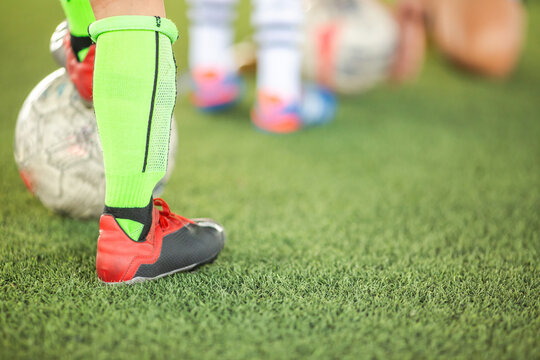 Selective focus to leg of youth football player standing on green artificial grass turf during a training session with blurry soccer balls in the background.