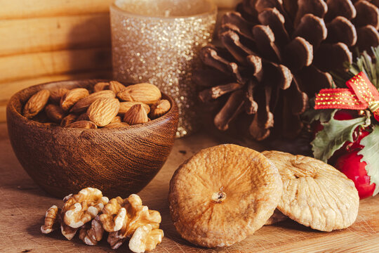 Festive holiday table displaying a wooden bowl of almonds, scattered walnuts, and dried figs, with a decorative pine cone and a sparkling candle creating a warm Christmas Eve atmosphere