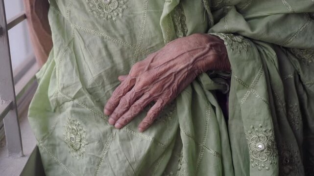 A hand of elder woman rests on a green garment in a room with good light