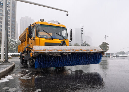 Yellow snowplow truck equipped with a wide blue brush clearing snow and slush from a city street during winter
