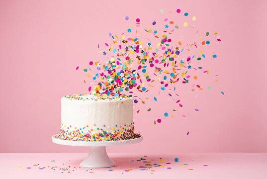 White birthday cake on a stand with a colorful confetti explosion against a pink background. Fun party celebration.
