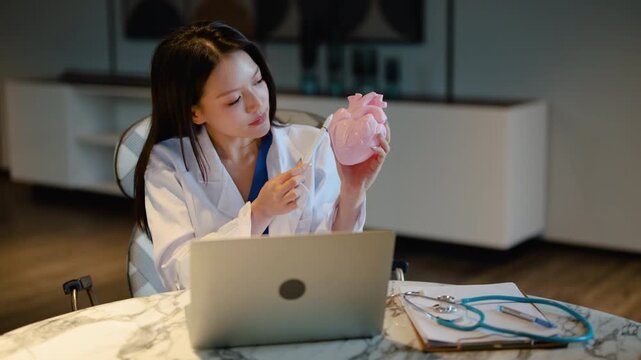 Asian female doctor holding pink heart model, explaining cardiology during virtual telemedicine consultation. Medical professional online video call for healthcare clinic education and expert advice.