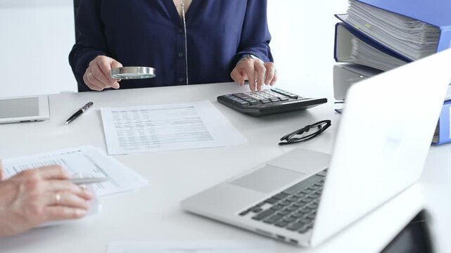 Female financial analysts examining documents, using magnifying glass and calculator while discussing corporate data with a male colleague. Audit and taxes in business