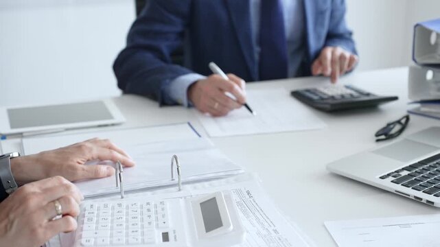 Two accountants are collaborating on a project, using a calculator and taking notes, surrounded by documents and technology in a busy office environment. Audit and taxes in business