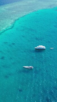 Aerial view turquoise reef with two boats anchored over shallow coral deepblue horizon sunlit water distant speedboat leaving faint wake tranquil atmosphere ideal for snorkeling, Maldives