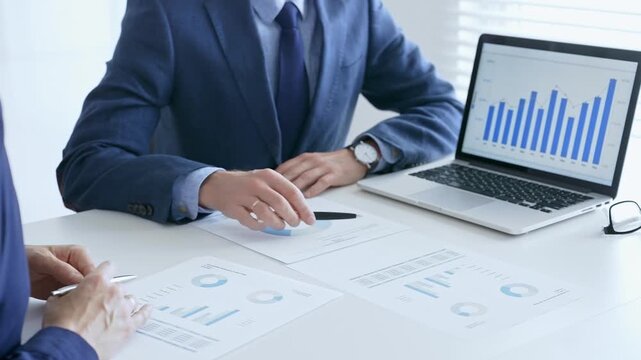 Business colleagues discussing financial data on a laptop and printed documents while analyzing charts and market trends. Business people and finance concept