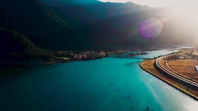 Aerial view of a turquoise river winding through mountainous valley with highway bridge and small village at golden hour in Tibet