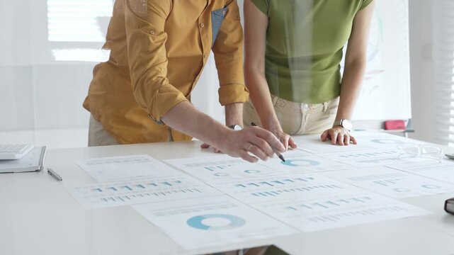 Business professionals analyzing financial reports, pointing at data points on charts and graphs lying across white desk surface