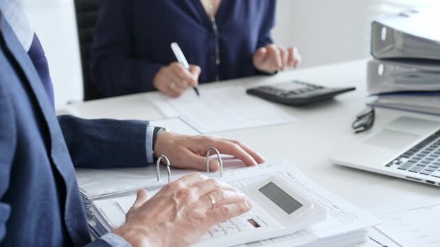 Two professional accountants collaborating in a modern office, calculating taxes and analyzing financial data with calculators, documents, and a laptop. Audit and taxes in business