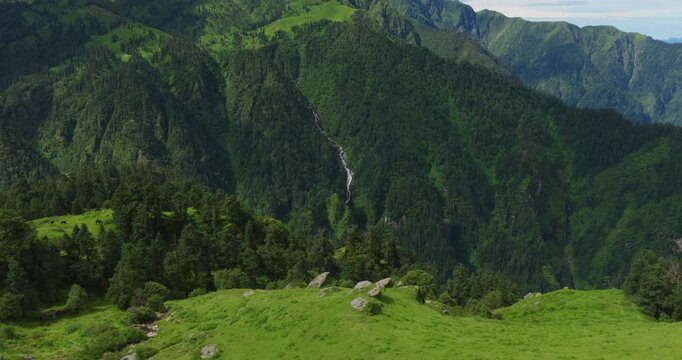 Badimalika, Bajura, Nepal, drone gliding forward over lush green alpine knolls and rocky outcrops toward expansive forested valleys and majestic Himalayan ridges under clear blue skies
