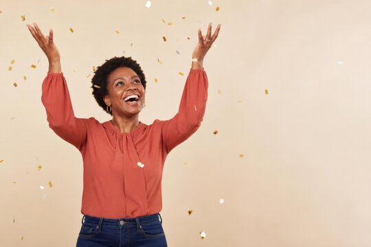 Smiling Black woman raising arms amid falling confetti against beige studio background