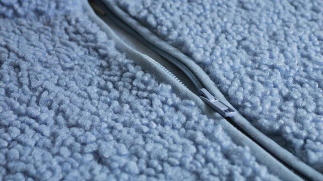 A close-up of a light blue sherpa fleece jacket zipper featuring a textured faux fur curly sheepskin wool, soft boucle fabric warmth, blue plastic slider, and fluffy textile structure details.