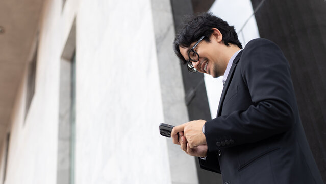 Smiling Asian businessman in a suit using a smartphone outdoors near a modern office building, checking messages, trading stocks, and managing mobile business communication online
