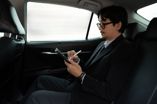Asian businessman using a digital tablet and stylus pen on the backseat of a moving car, working productively during commute, executive lifestyle, modern mobility and wireless technology concept