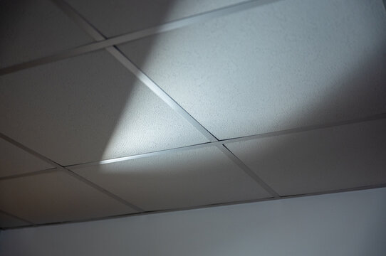Suspended acoustic tile ceiling illuminated by a beam of light, creating dramatic geometric shadows and an abstract architectural pattern in a modern interior.