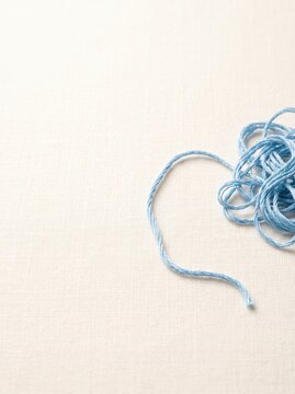 Loose blue thread rests on white fabric. A tangled coil sits at the right edge with a long strand crossing the textured surface and copy space. Craft, sewing, and handmade design concept.