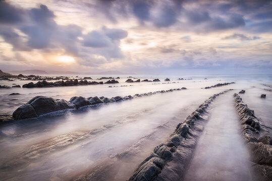 Sunset over Barrika beach with rocky flysch formations and dramatic waves crashing under a golden orange sky, Biscay, Basque Country, Spain
