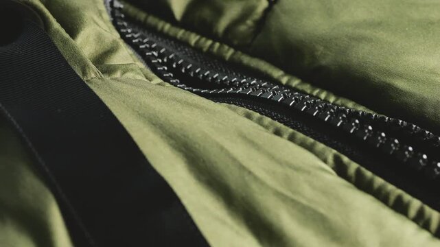 A close-up of a green quilted jacket zipper mechanism reveals olive puffer coat texture, fabric folds, a heavy plastic zipper, a black adjustment strap, a shiny metal grommet, and sewing craftsmanship