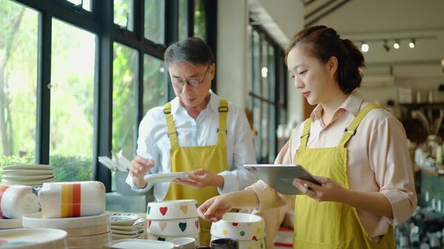 Asian male and female pottery shop owners wearing yellow aprons checking ceramic plates and stock inventory with a digital tablet. Small business retail teamwork and modern entrepreneurship concept.