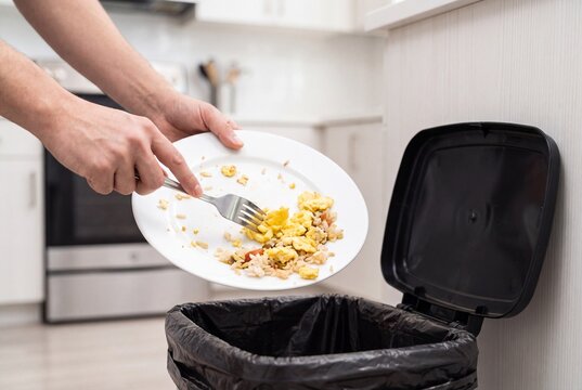 Person Scraping Food Leftovers from Plate into Trash Can in Kitchen