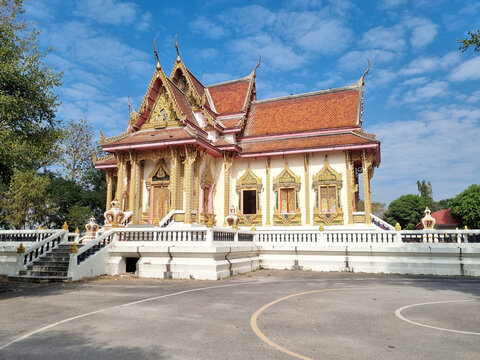 Traditional Thai Buddhist temple with ornate golden architecture and orange tiled roof under a bright blue sky. Beautiful religious building in Thailand featuring decorative gables and white walls.