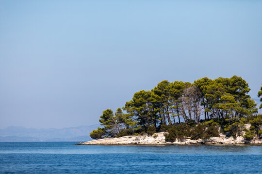 A panoramic view of a small, tree-covered island surrounded by the crystal-clear blue waters of the Mediterranean Sea. Lush green vegetation contrasts with the deep blue sea.