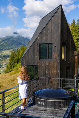 Woman in Bathrobe on Terrace Beside Outdoor Hot Tub. Woman in a white bathrobe stands on a mountain terrace beside a round outdoor hot tub and dark wooden cabin under a blue sky. © Iryna