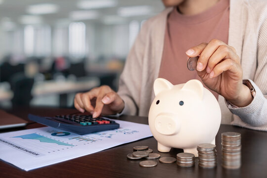 Save money and budget concept with a woman putting a coin into a piggy bank. Using a calculator and analyzing financial charts on her desk.