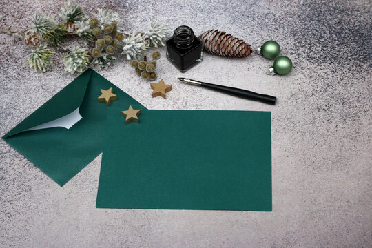 Elegant Christmas mockup featuring a blank green greeting card, an envelope, a fountain pen, an inkwell, and festive decorations on a concrete background