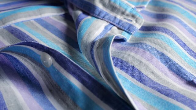 A close-up of a striped linen shirt collar and buttons features alternating blue, grey, and white vertical stripes, natural light texture, pearl buttons, and casual male garment details.