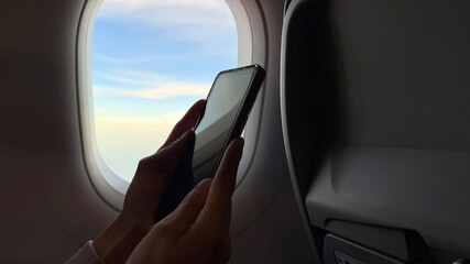 Obraz premium Close up of traveler hands holding and using a mobile smartphone inside an airplane cabin next to a window with a scenic blue sky view, digital flight booking and inflight connection concept