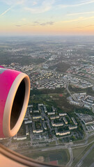 Obraz premium Aerial view of a city landscape with houses and streets through an airplane window next to a pink jet engine during flight, commercial aviation tourism and travel wanderlust concept